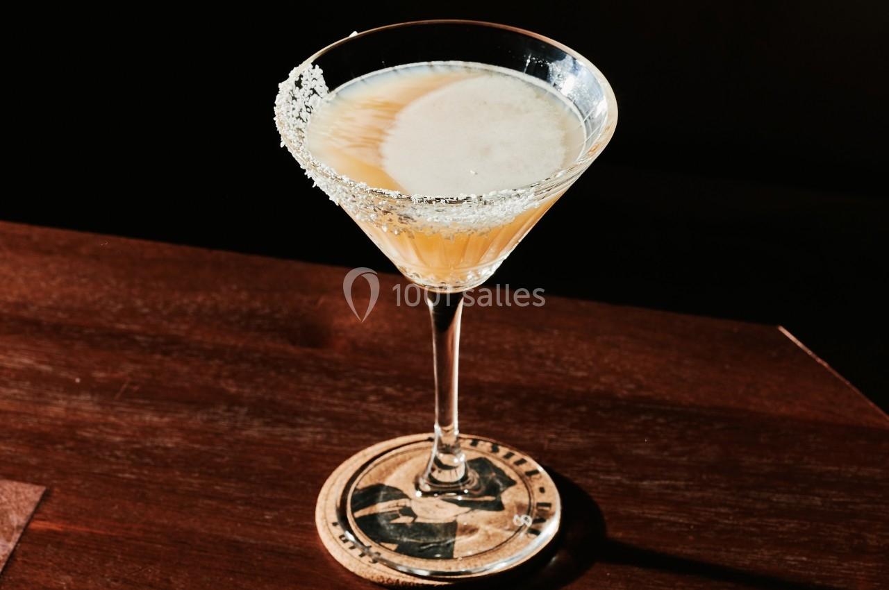 Verre à cocktail avec bord salé, posé sur un sous-verre en bois, éclairé par une lumière tamisée.