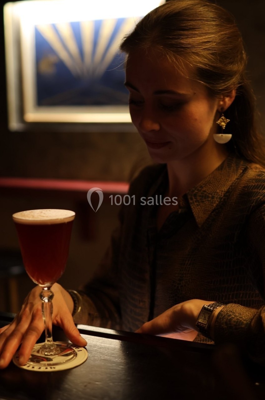 Une femme assise dans un bar sombre tient un menu près d'un cocktail rouge servi dans un verre élégant.