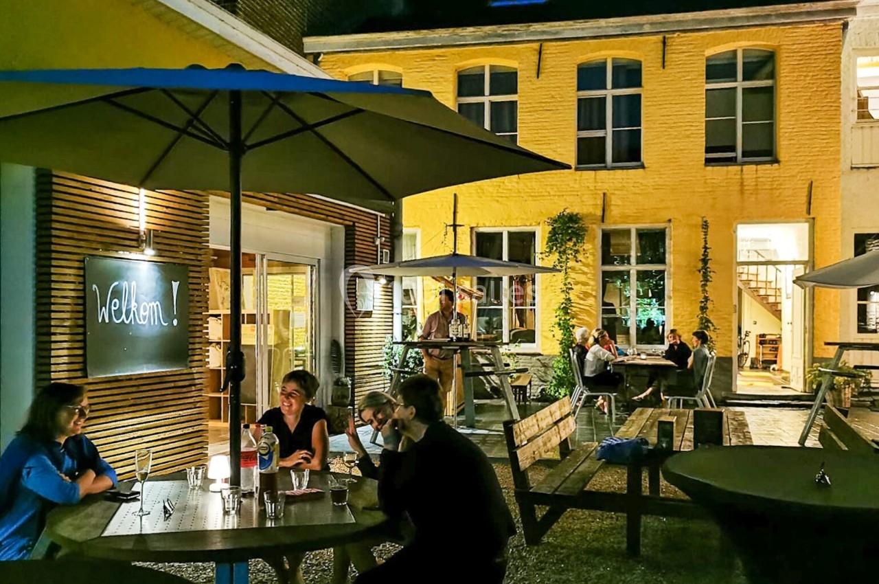 Terrasse extérieure éclairée en soirée, avec des personnes assises autour de tables sous des parasols devant un bâtiment…