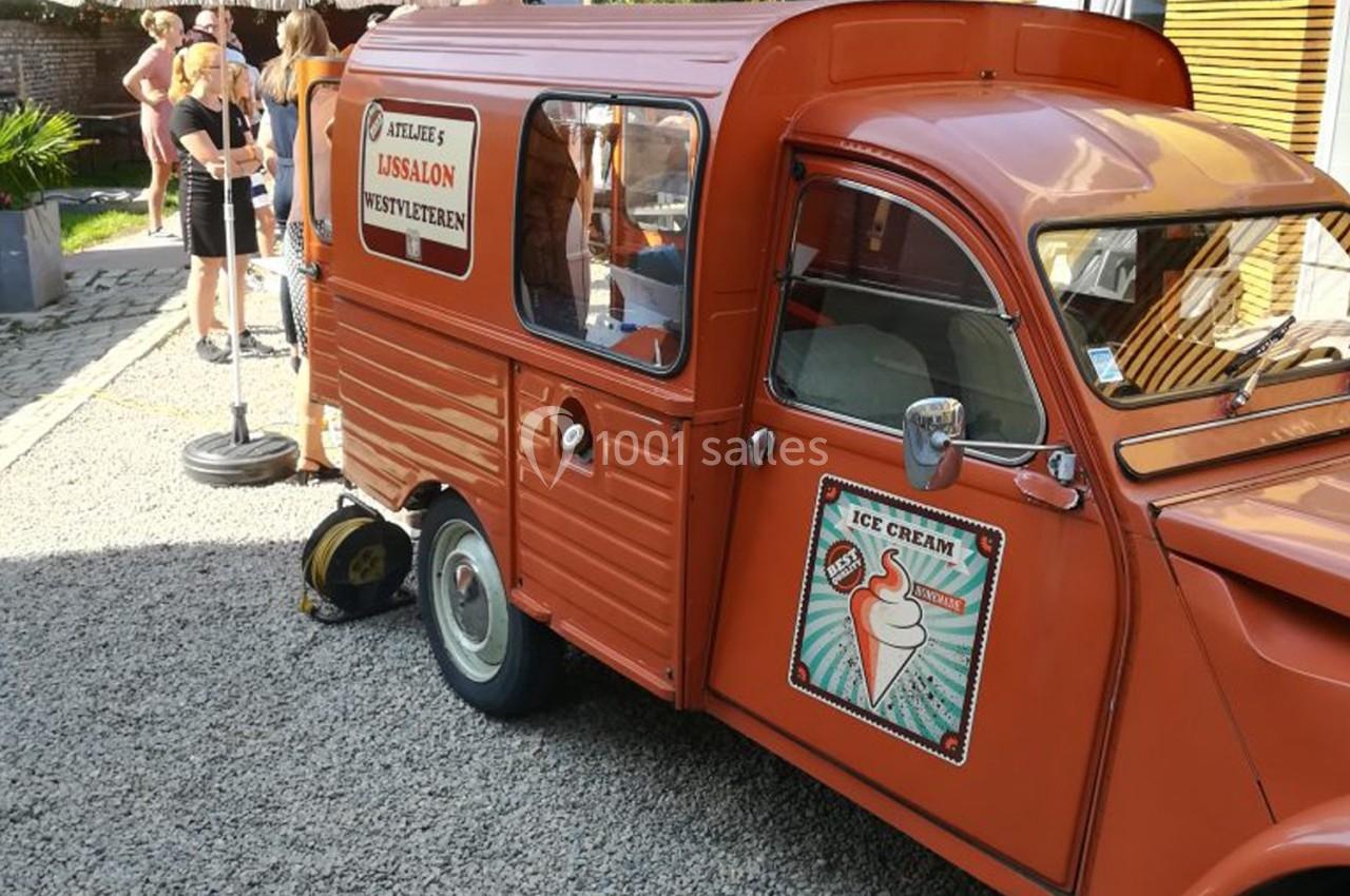 Camionnette vintage orange transformée en stand de glaces, stationnée sur un sol gravillonné près de personnes.