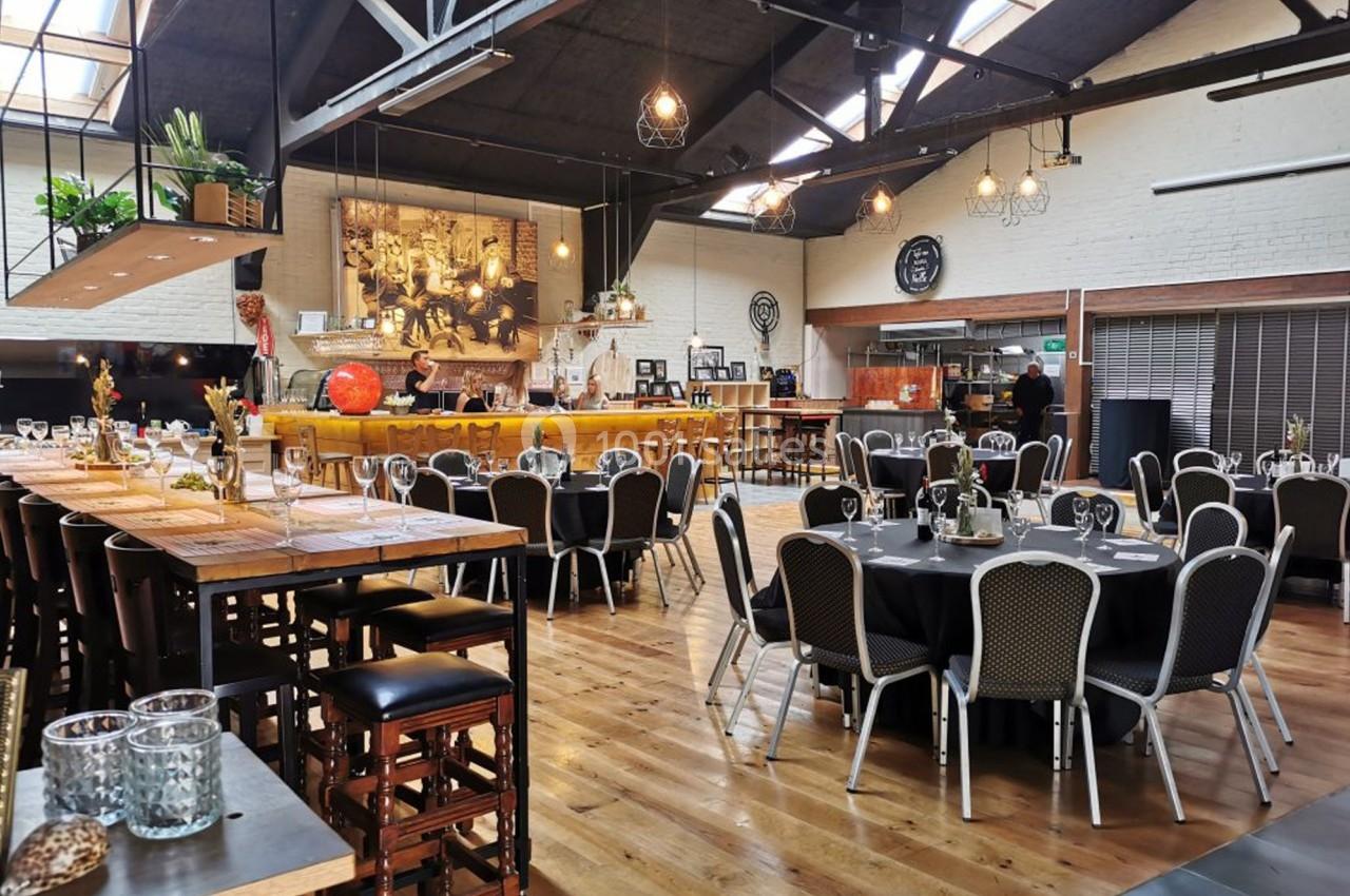 Salle de réception lumineuse avec tables dressées, chaises noires et décoration industrielle moderne.