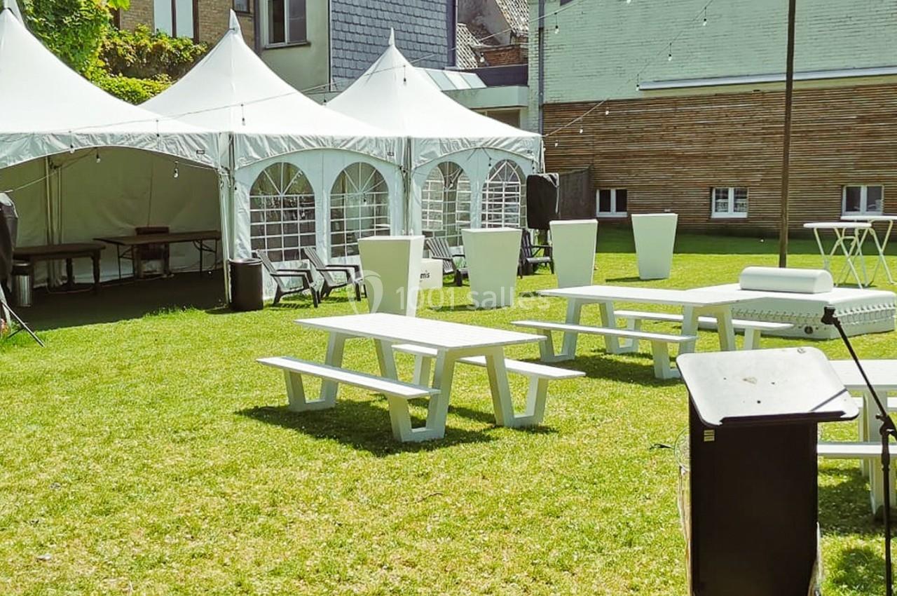 Tables de pique-nique blanches et tentes installées sur une pelouse, entourées de bâtiments en arrière-plan.