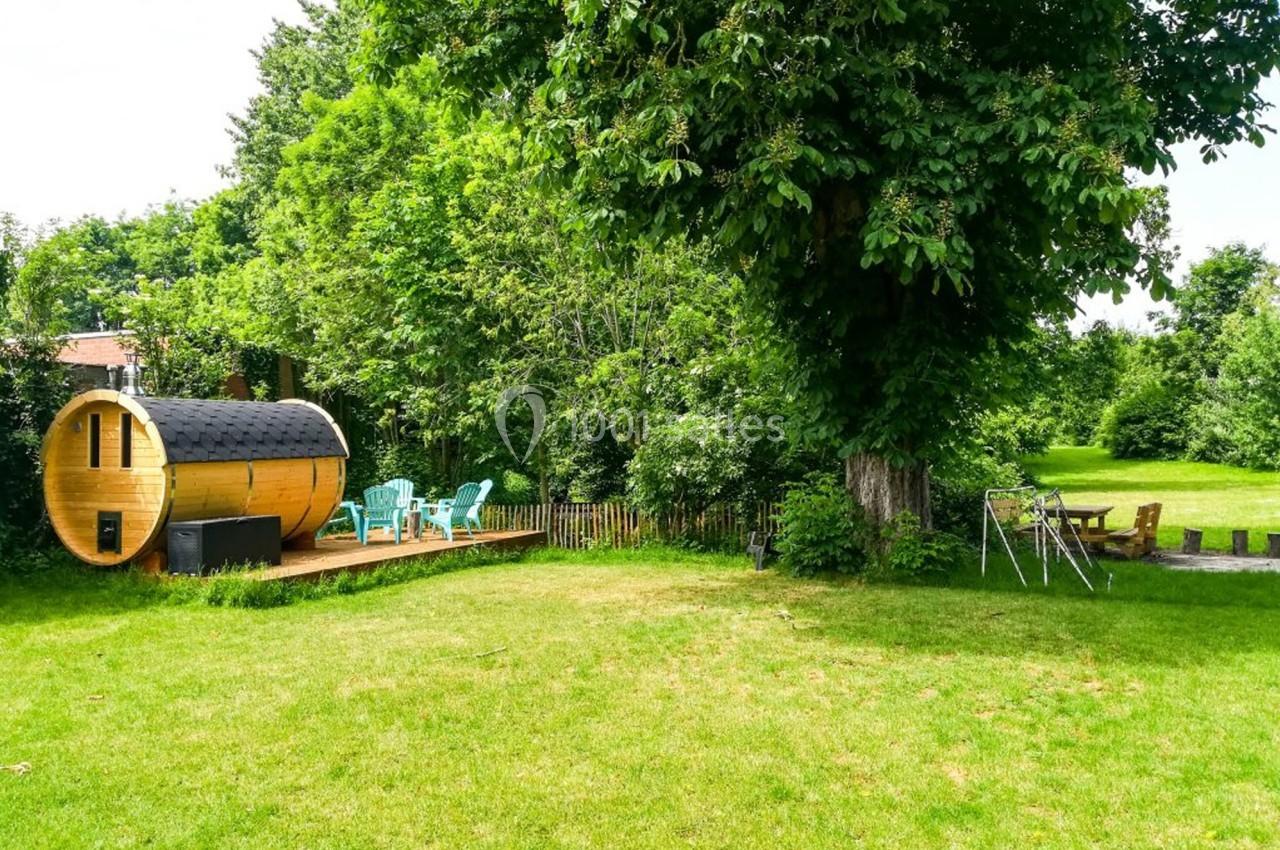 Jardin verdoyant avec sauna en bois, chaises turquoise sur une terrasse et balançoire près d'un grand arbre.