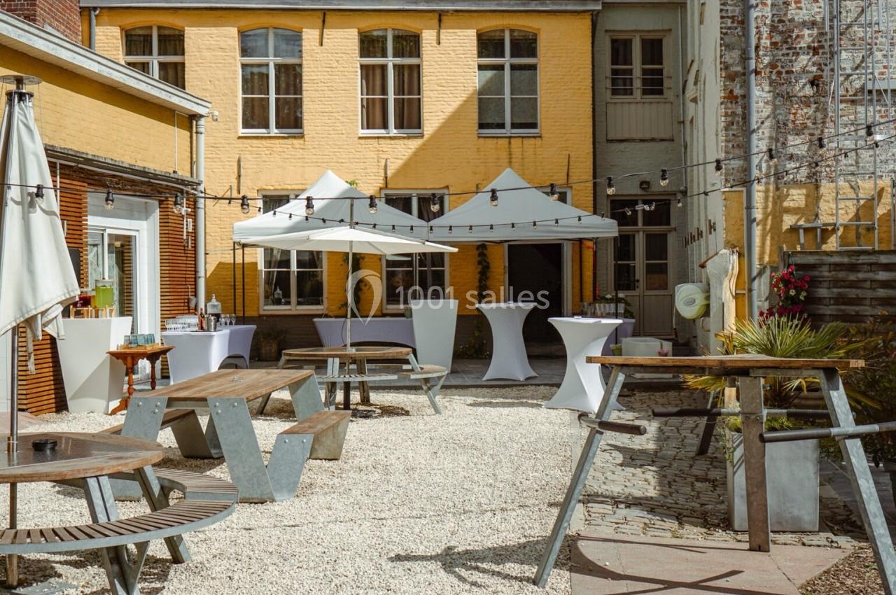 Cour intérieure avec tables en bois, parasols blancs et guirlandes lumineuses, entourée de bâtiments en briques jaunes.