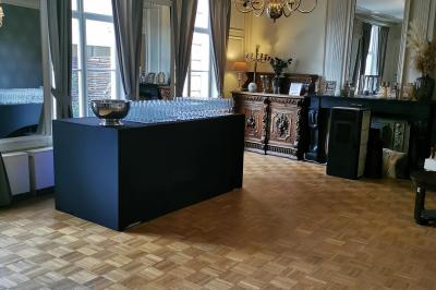 Salle élégante avec parquet, comptoir noir, lustre central et grandes fenêtres encadrées de rideaux.