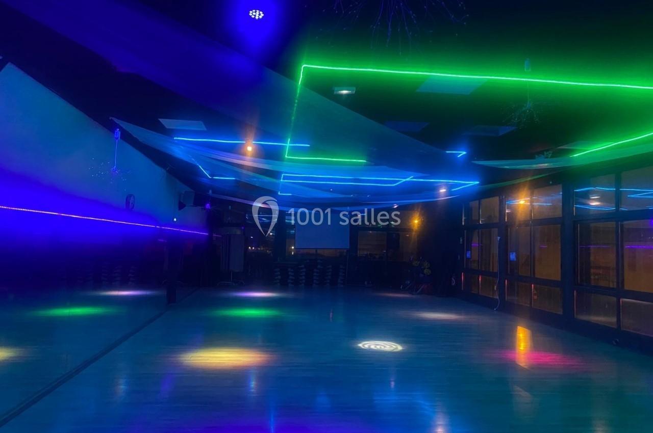 Salle sombre avec un sol brillant, éclairée par des néons colorés et des reflets lumineux au plafond.