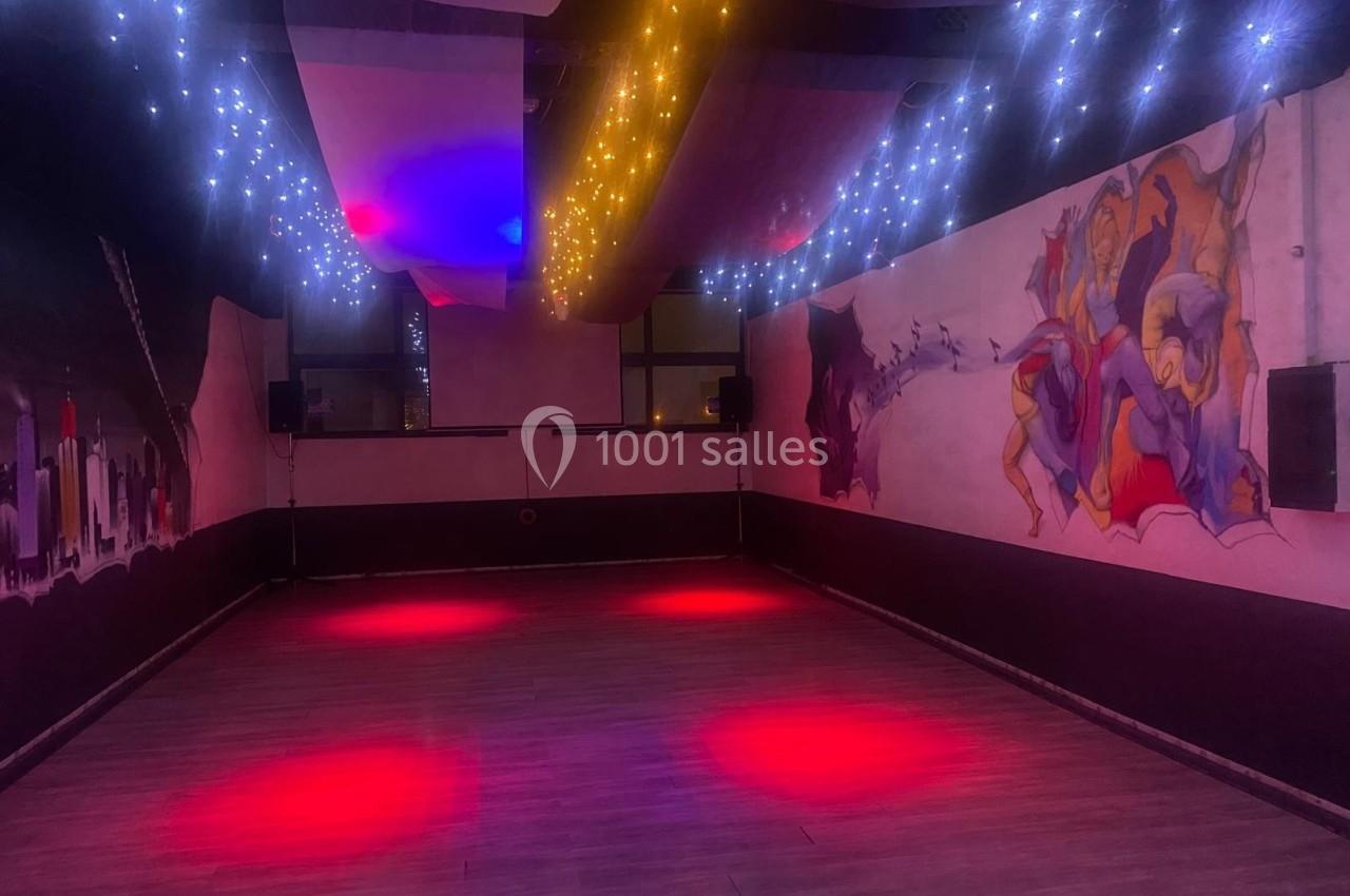 Salle sombre avec parquet, éclairée par des lumières rouges et guirlandes lumineuses au plafond, murs décorés de fresques.