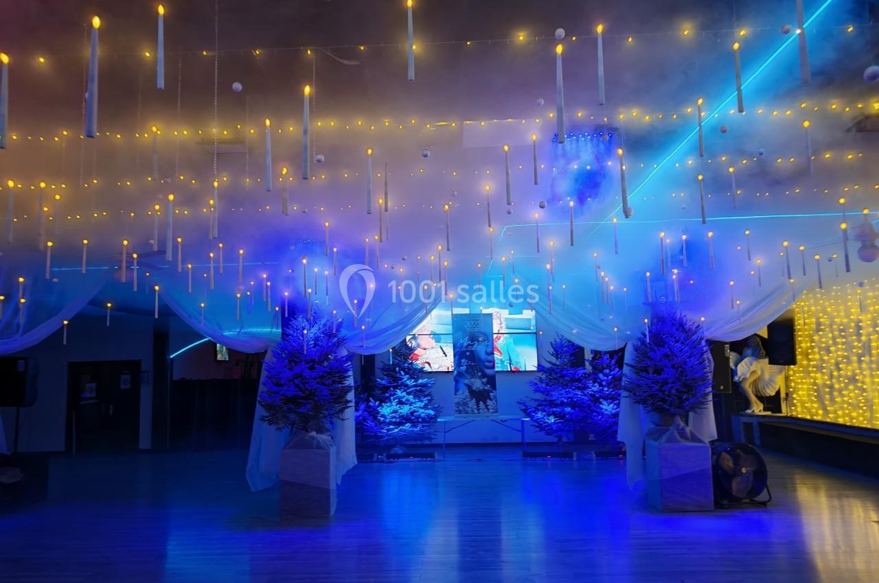 Salle décorée avec des guirlandes lumineuses suspendues, des sapins artificiels et un éclairage bleu tamisé.