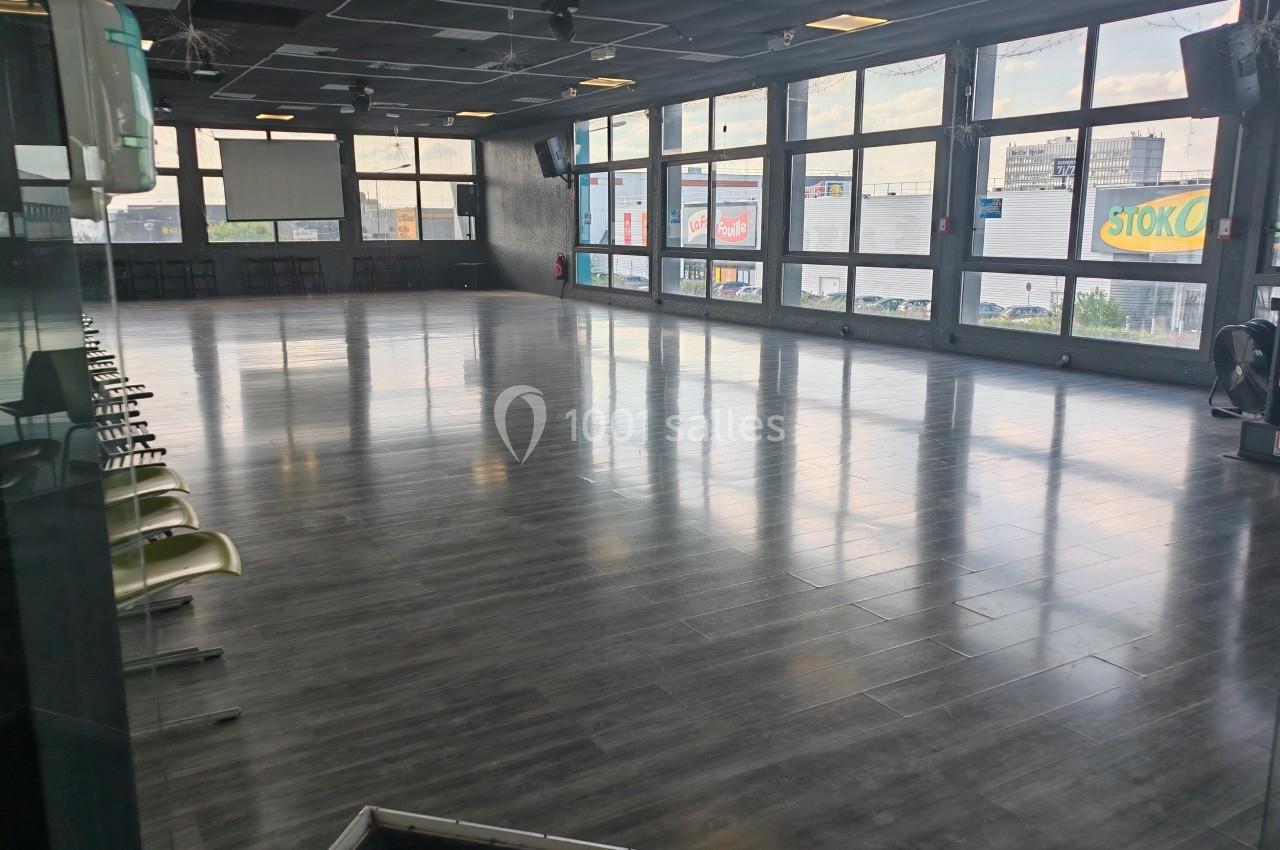 Salle spacieuse avec parquet sombre, grandes baies vitrées et chaises alignées sur le côté.