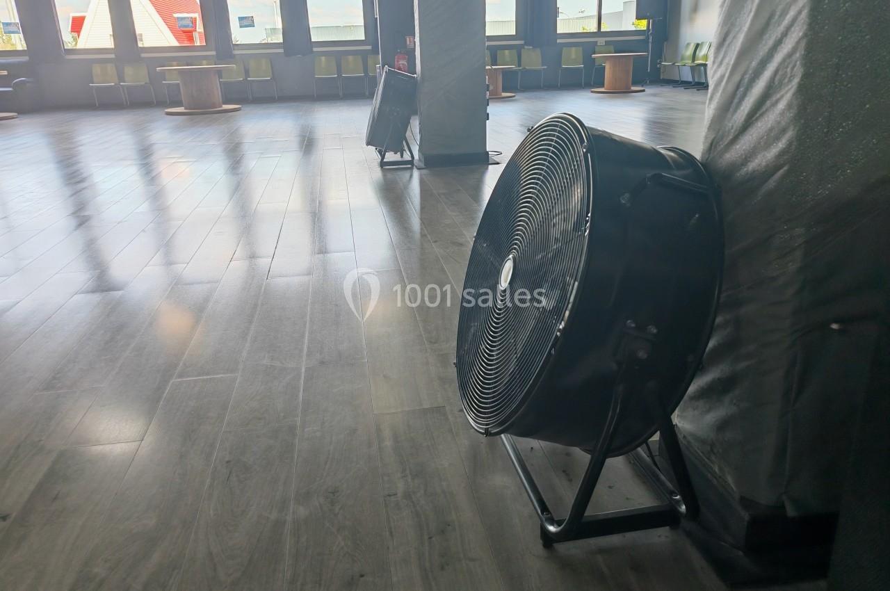 Un grand ventilateur noir posé au sol dans une pièce lumineuse avec un sol en parquet et des fenêtres en arrière-plan.