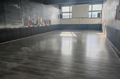 Salle vide avec un sol en parquet sombre, éclairée par la lumière naturelle entrant par de grandes fenêtres.