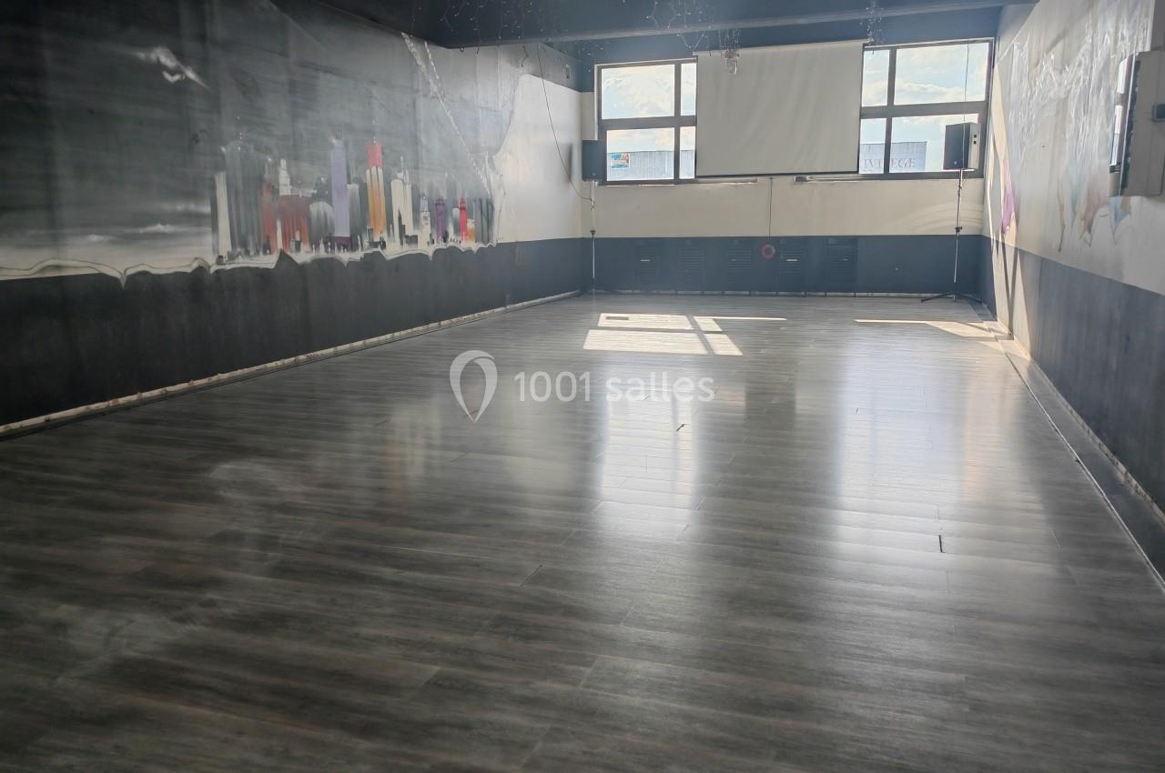 Salle vide avec un sol en parquet sombre, éclairée par la lumière naturelle entrant par de grandes fenêtres.