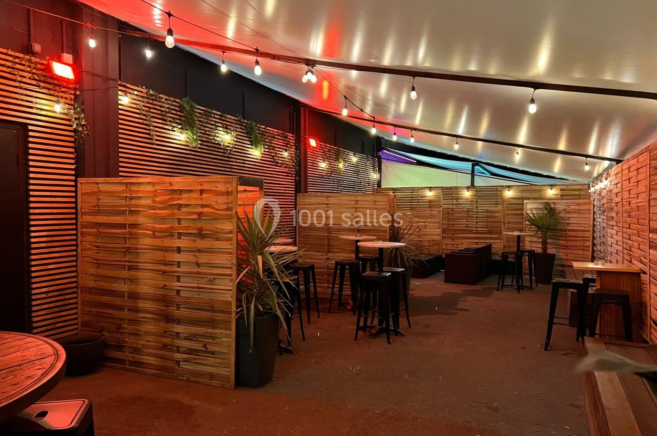 Terrasse extérieure couverte avec éclairage suspendu, séparations en bois, tables hautes et plantes décoratives.