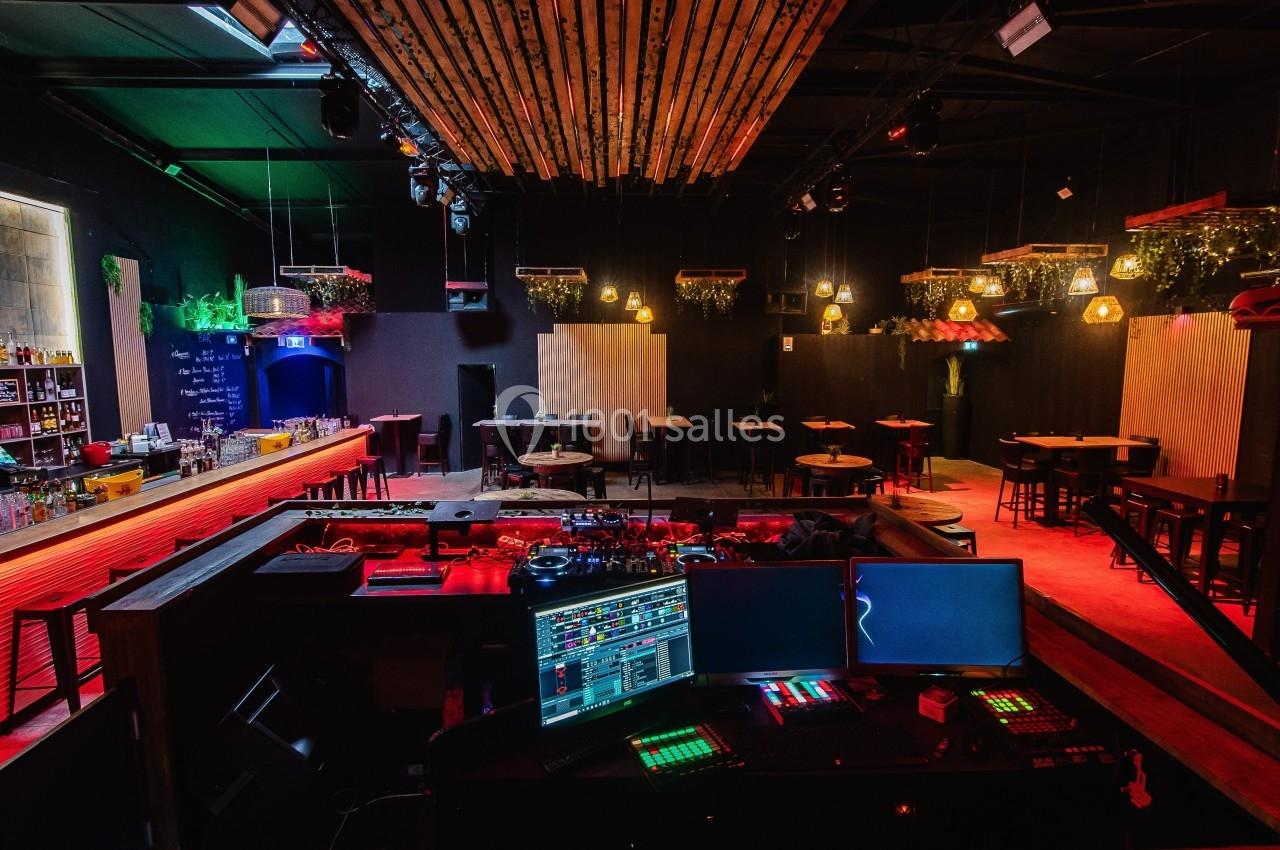 Salle de bar avec éclairage tamisé, tables, bar à gauche et espace DJ avec équipements électroniques au premier plan.