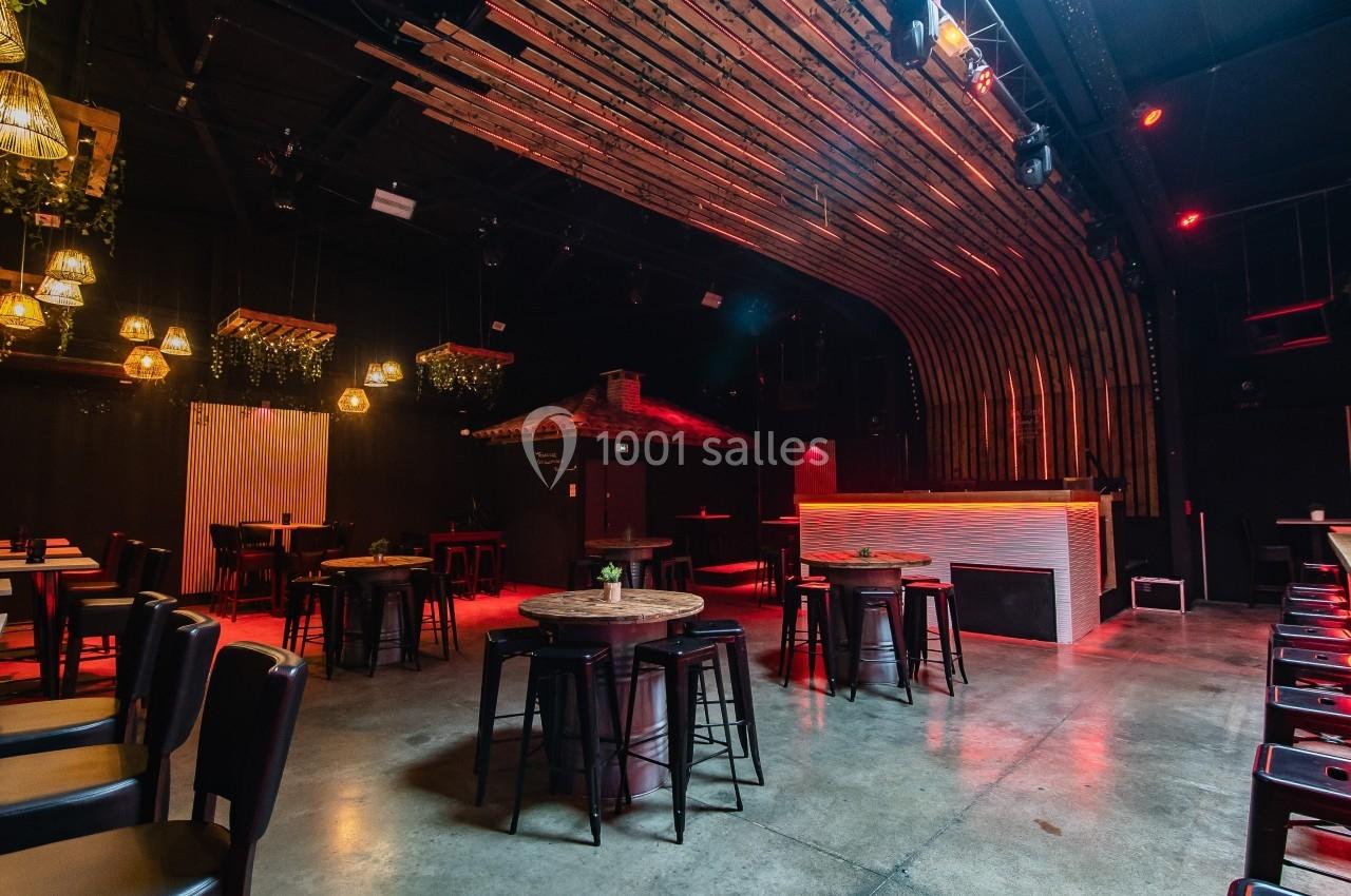 Salle de bar moderne avec tables hautes, tabourets, éclairage tamisé et décorations suspendues en bois et plantes.