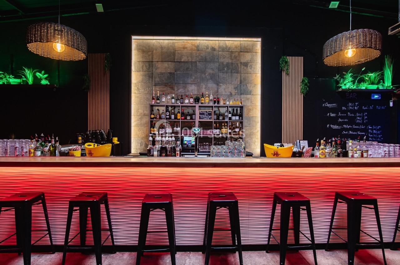 Bar moderne avec comptoir éclairé en rouge, tabourets noirs, étagères de boissons et décoration végétale.