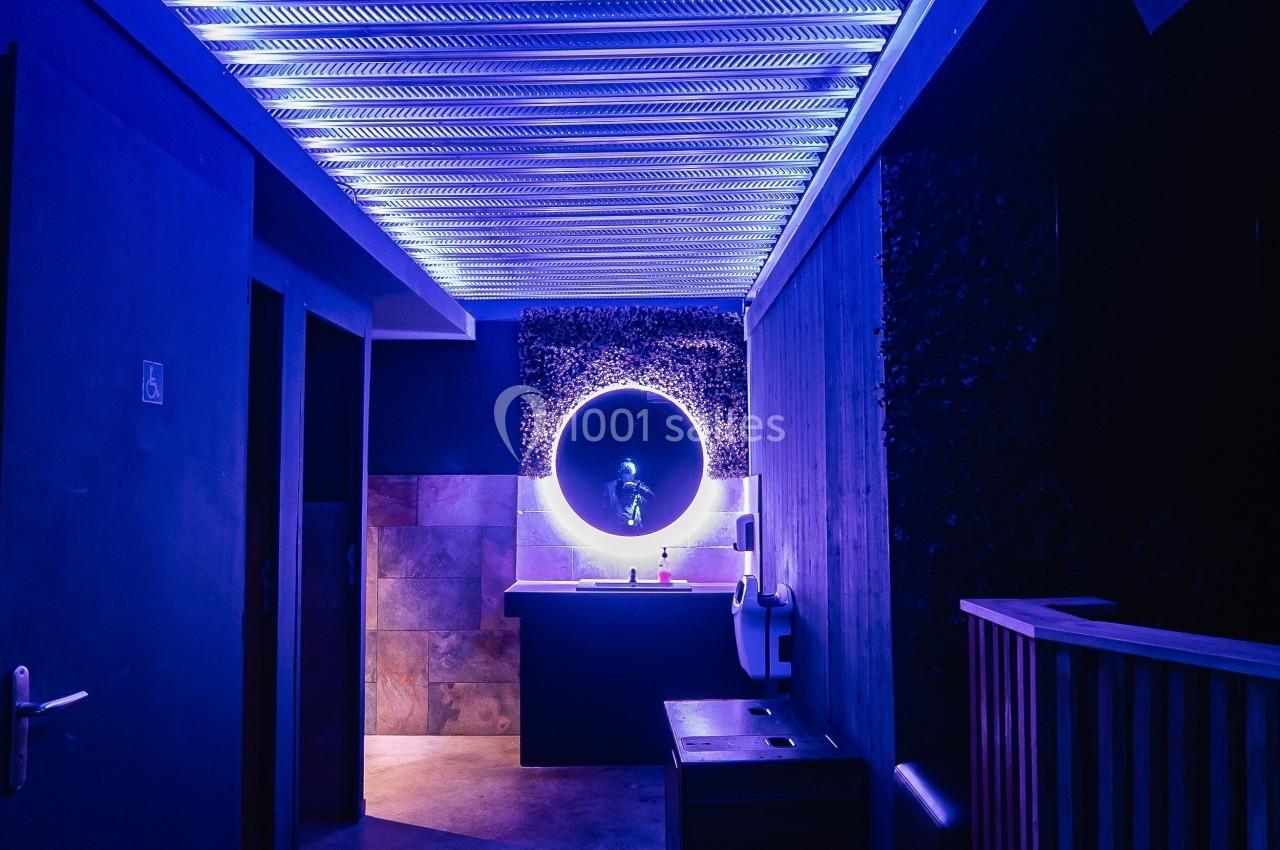 Couloir sombre avec éclairage violet, menant à un lavabo éclairé par un miroir rond lumineux.