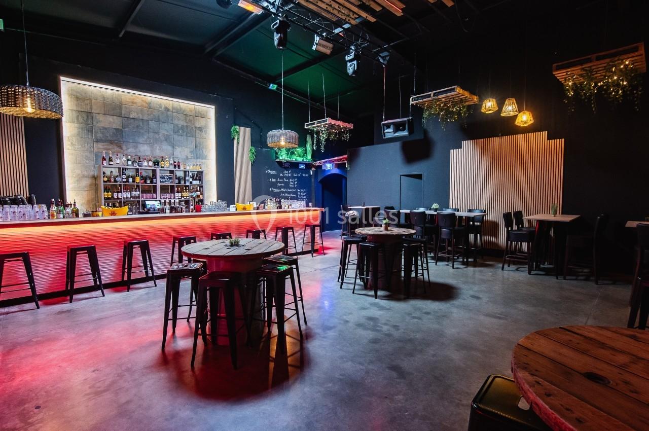 Salle de bar moderne avec comptoir lumineux, tables hautes en bois, chaises noires et éclairage tamisé.