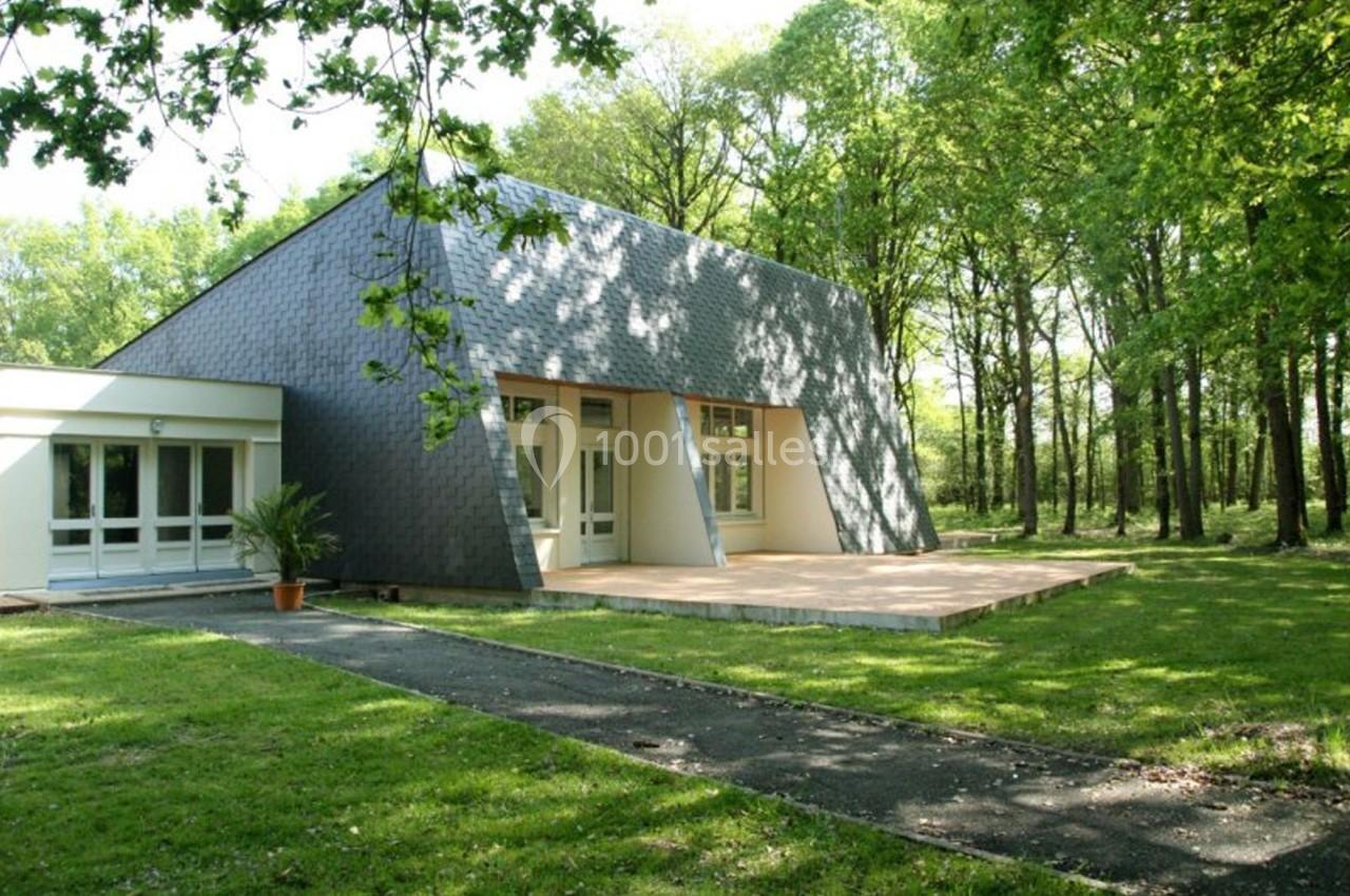 Bâtiment moderne avec façade en ardoise, grandes baies vitrées et terrasse en bois, entouré d'arbres et de pelouse.