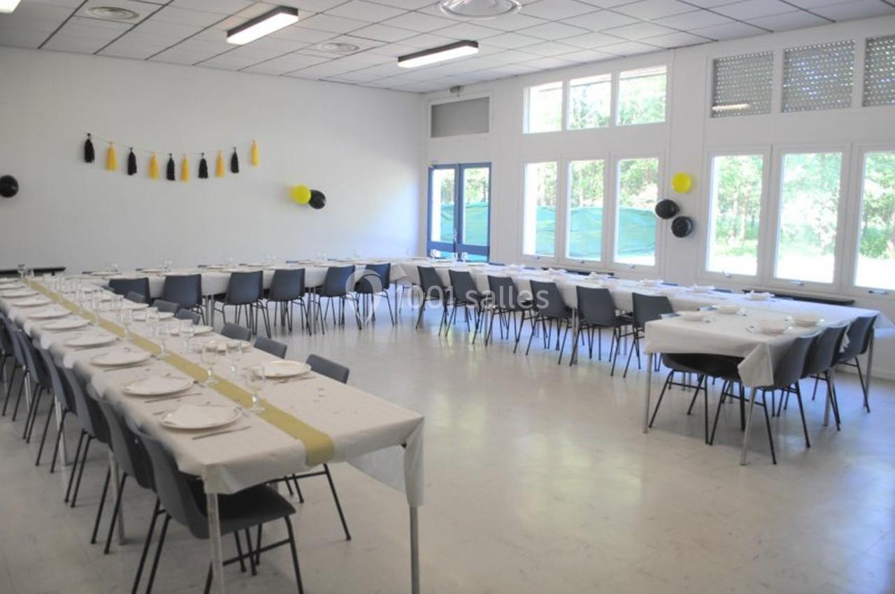 Salle lumineuse aménagée avec des tables alignées et décorées pour un événement, vue sur des fenêtres donnant sur l…
