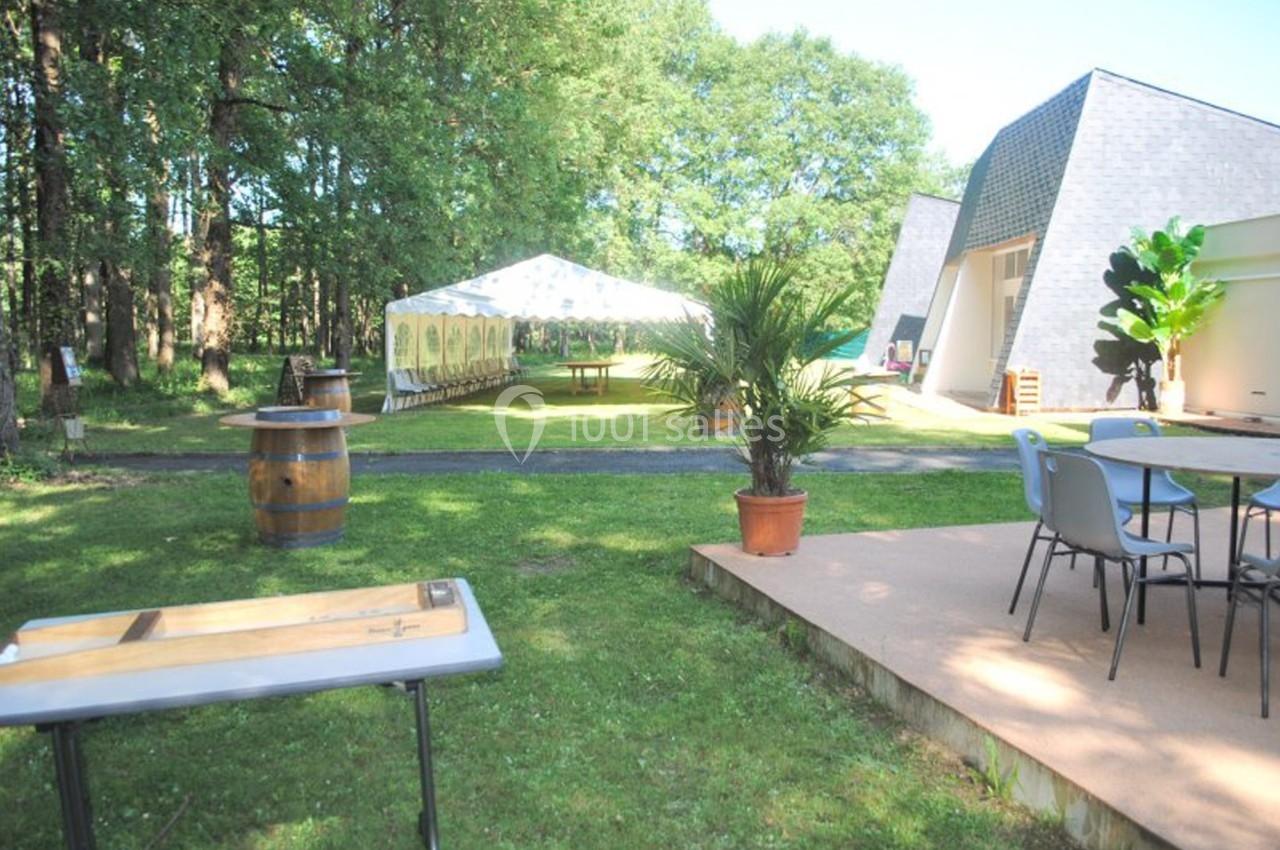 Espace extérieur avec pelouse, tables, chaises, une tente blanche et un bâtiment moderne entouré d'arbres.