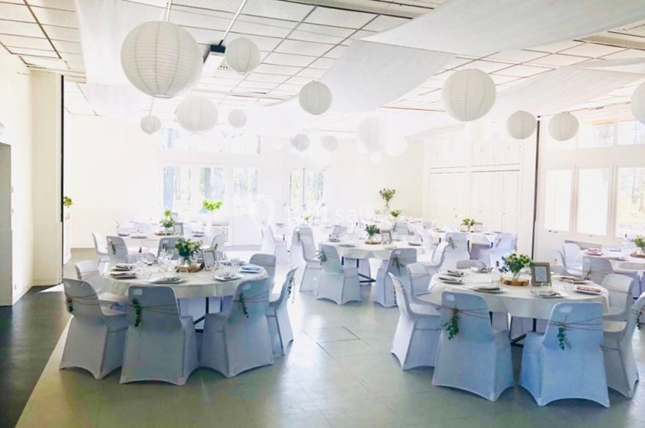 Salle de réception lumineuse avec tables rondes dressées, nappes blanches et lanternes suspendues au plafond.