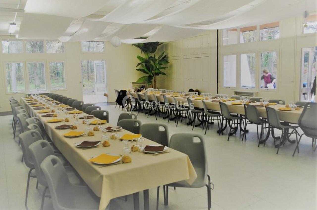 Salle de réception avec deux longues tables dressées, nappes beige, assiettes jaunes et chaises grises, éclairée par la…