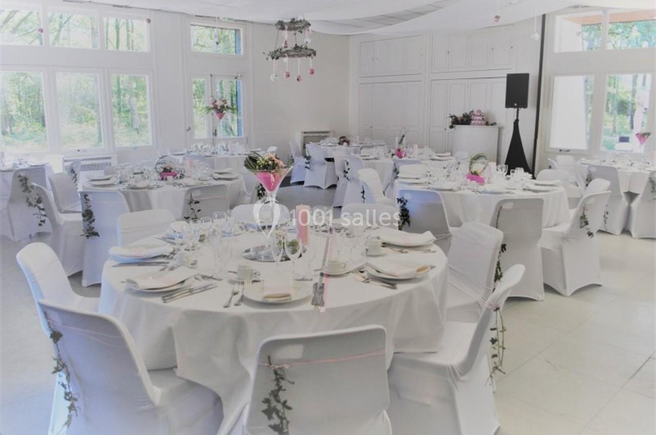 Salle de réception décorée pour un mariage, avec des tables rondes dressées et des chaises blanches ornées de guirlandes.