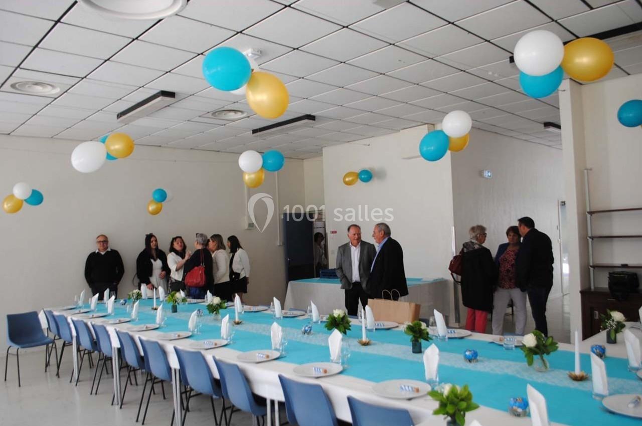Salle décorée avec des ballons bleus, blancs et dorés, tables dressées et groupe de personnes discutant debout.