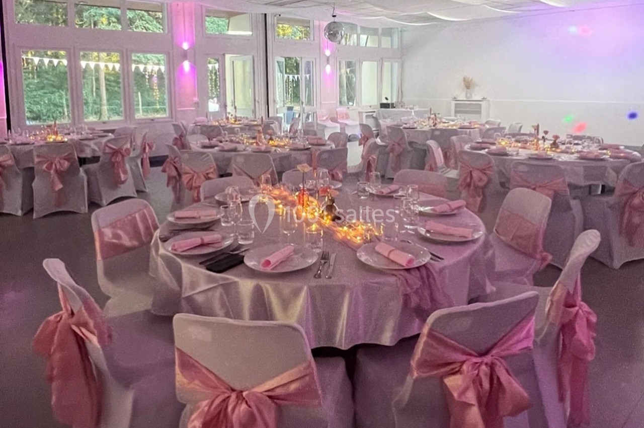 Salle de réception décorée avec des tables rondes couvertes de nappes blanches, ornées de rubans roses et de lumières…