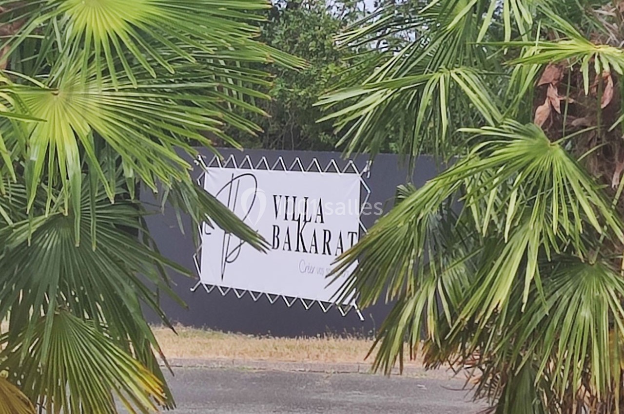 Panneau ’Villa Bakarat’ entouré de feuillages de palmiers, situé devant une clôture noire.