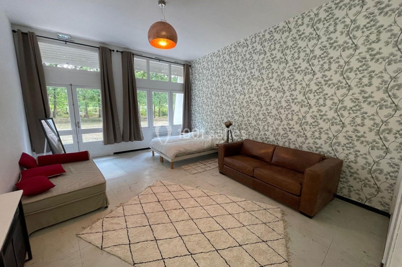 Salon lumineux avec canapé, lit, tapis beige, murs à motifs floraux et grandes fenêtres donnant sur un espace vert.