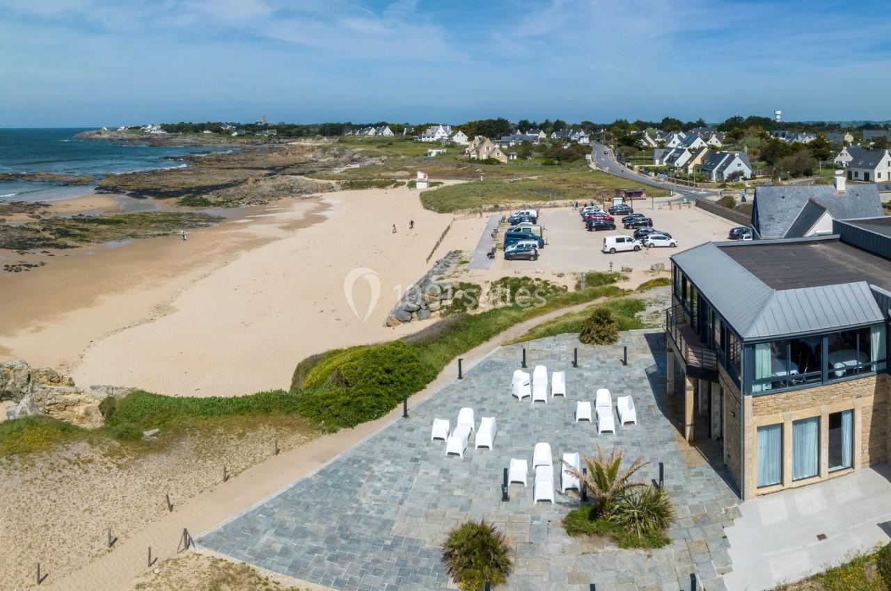 Vue aérienne d'une plage avec un parking, des maisons en arrière-plan et une terrasse aménagée au premier plan.