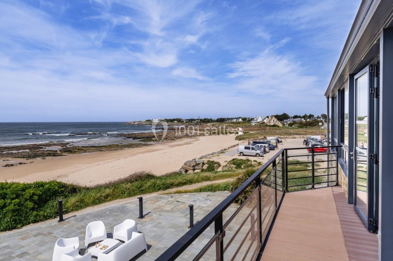 Vue depuis une terrasse avec mobilier extérieur, donnant sur une plage, des rochers et un ciel partiellement nuageux.