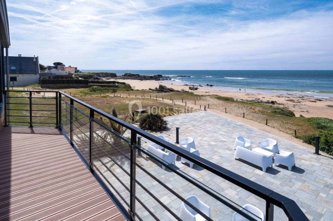 Vue depuis une terrasse surplombant une plage, avec des chaises blanches sur une esplanade ensoleillée.