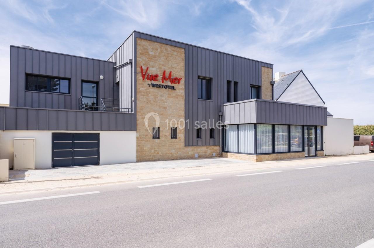 Façade moderne d'un bâtiment en pierre et métal avec une enseigne rouge visible, situé au bord d'une route.