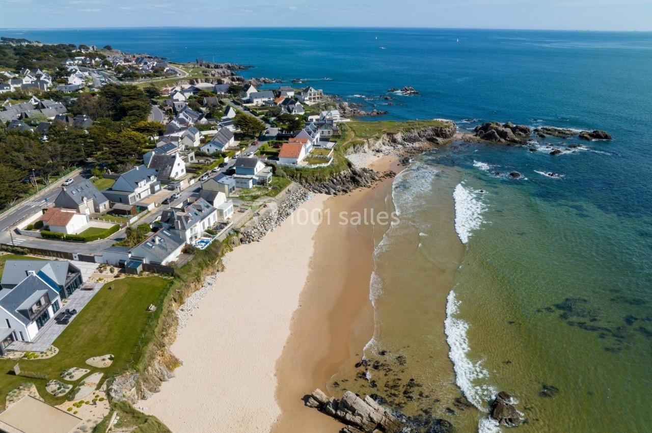 Vue aérienne d'une plage bordée de maisons, avec une mer calme et un littoral rocheux sous un ciel dégagé.