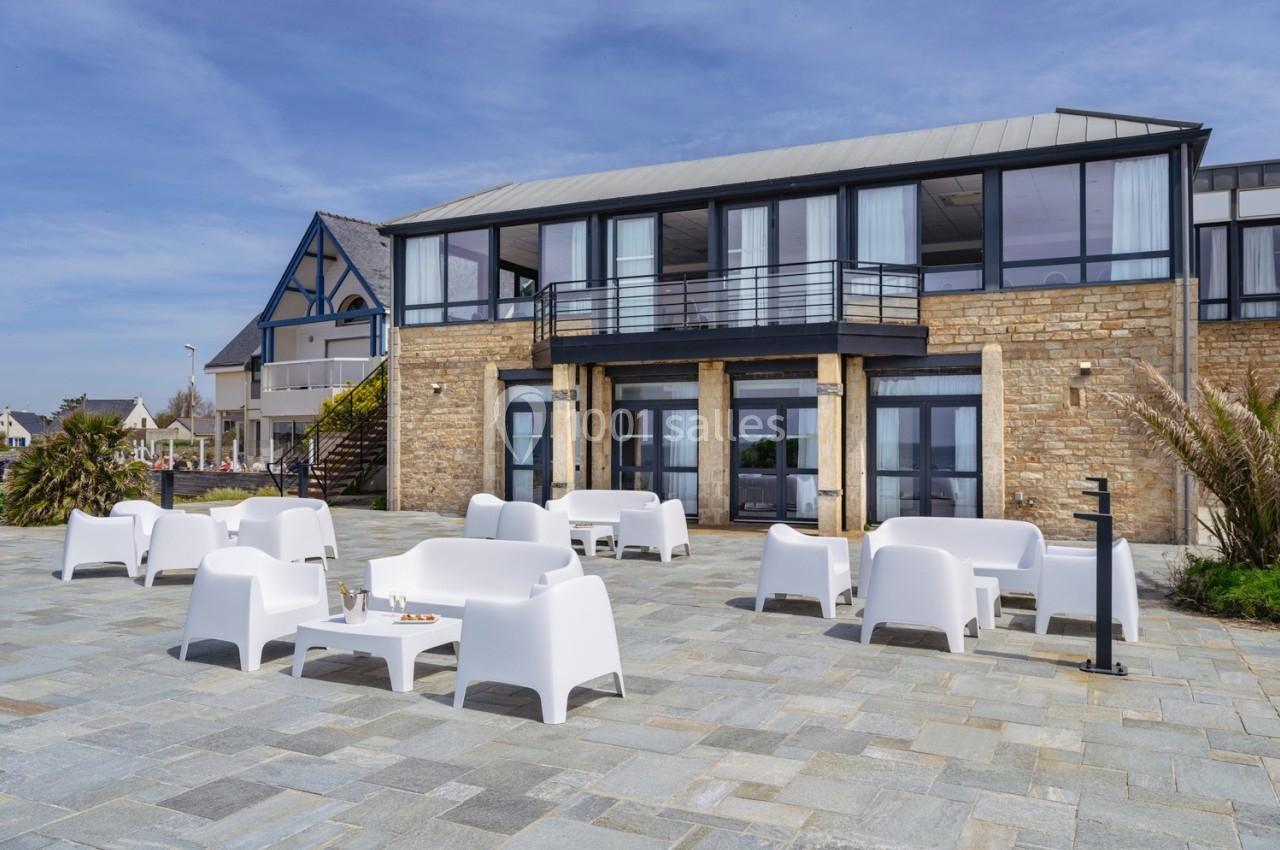 Terrasse en pierre avec mobilier blanc moderne devant un bâtiment à façade en pierre et grandes baies vitrées.