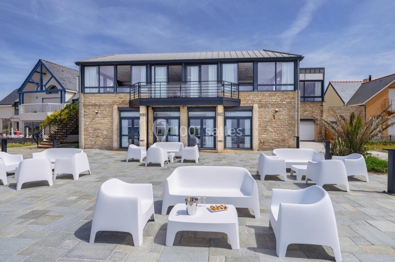 Façade d'un bâtiment moderne avec terrasse aménagée de mobilier blanc sur un sol en pierre, sous un ciel dégagé.