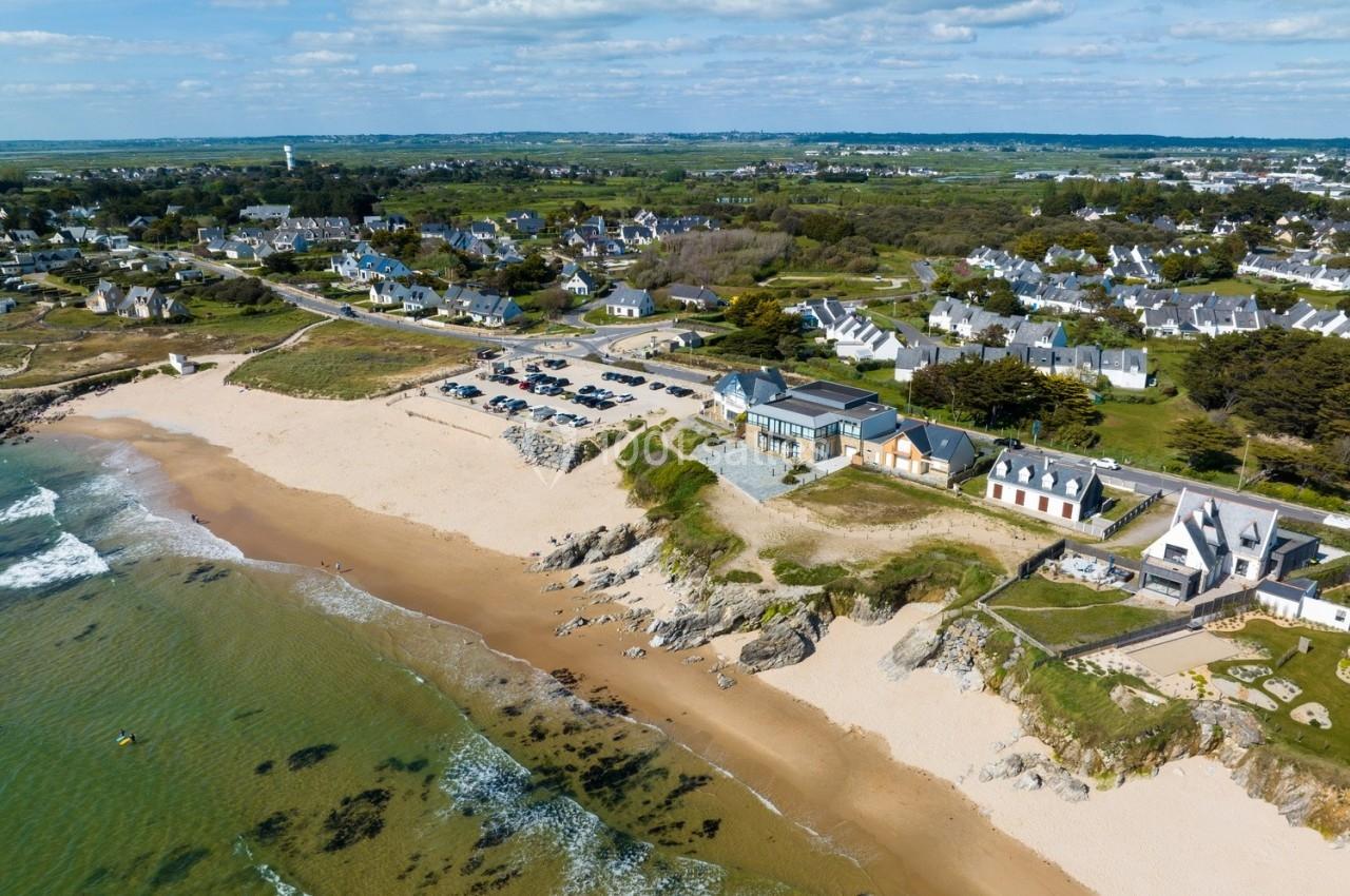 Vue aérienne d'une plage bordée de maisons, d'un parking et d'une végétation côtière sous un ciel partiellement nuageux.