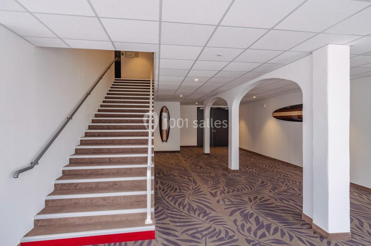 Escalier intérieur avec rampe, moquette à motifs au sol, murs blancs et décoration murale en bois.