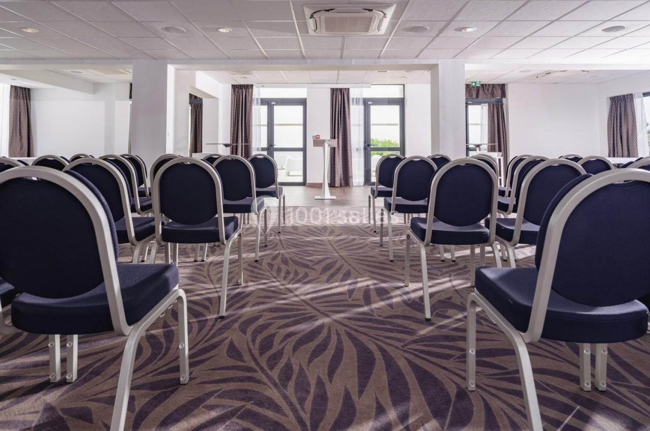 Salle de conférence vide avec des rangées de chaises noires et une moquette à motifs, éclairée par des fenêtres.