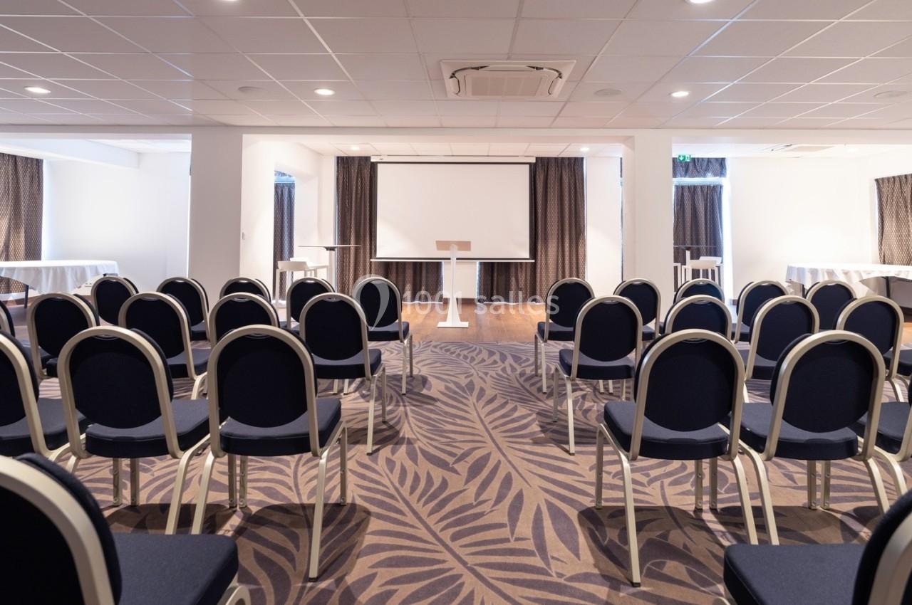 Salle de conférence avec des rangées de chaises bleues, un podium et un écran blanc au fond.