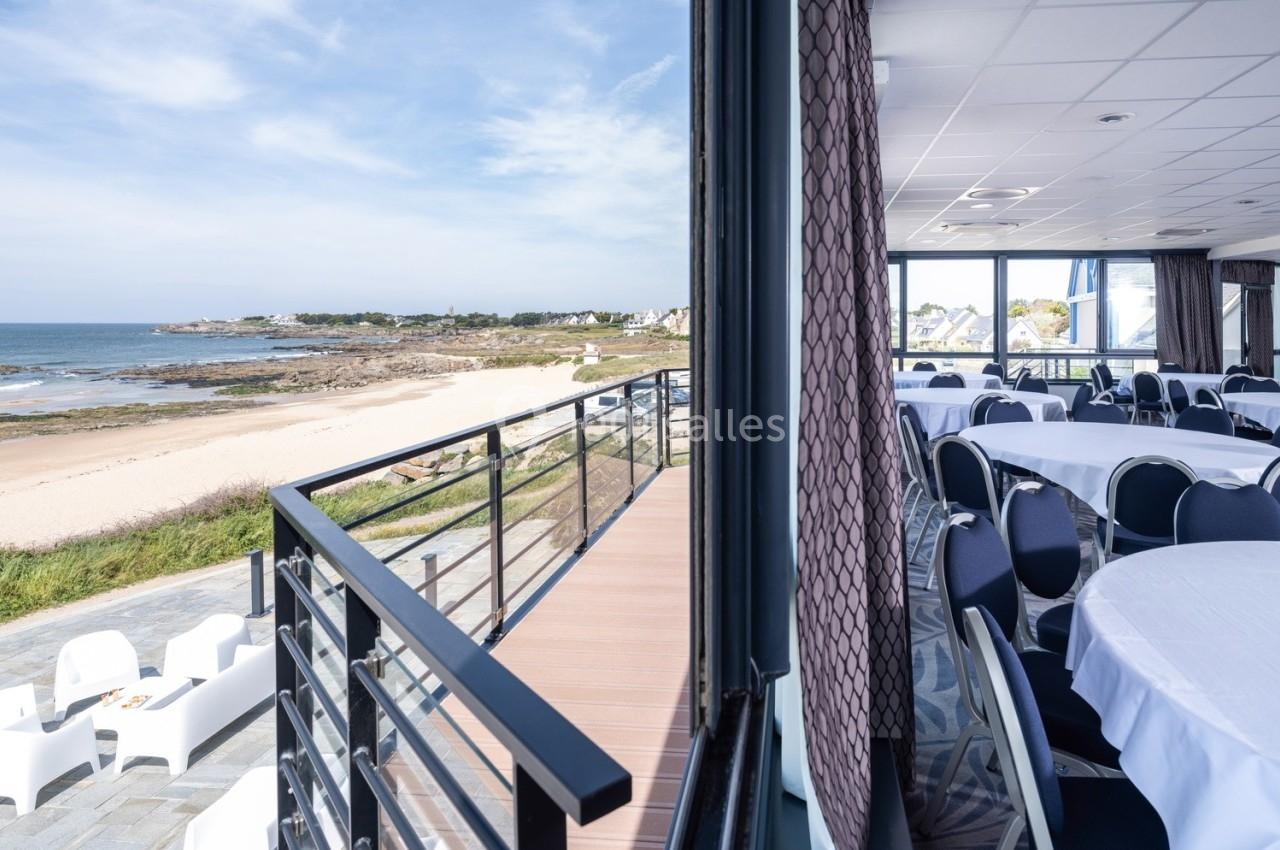 Vue d'une salle de réception lumineuse avec des tables rondes, donnant sur une terrasse et une plage bordée de rochers.