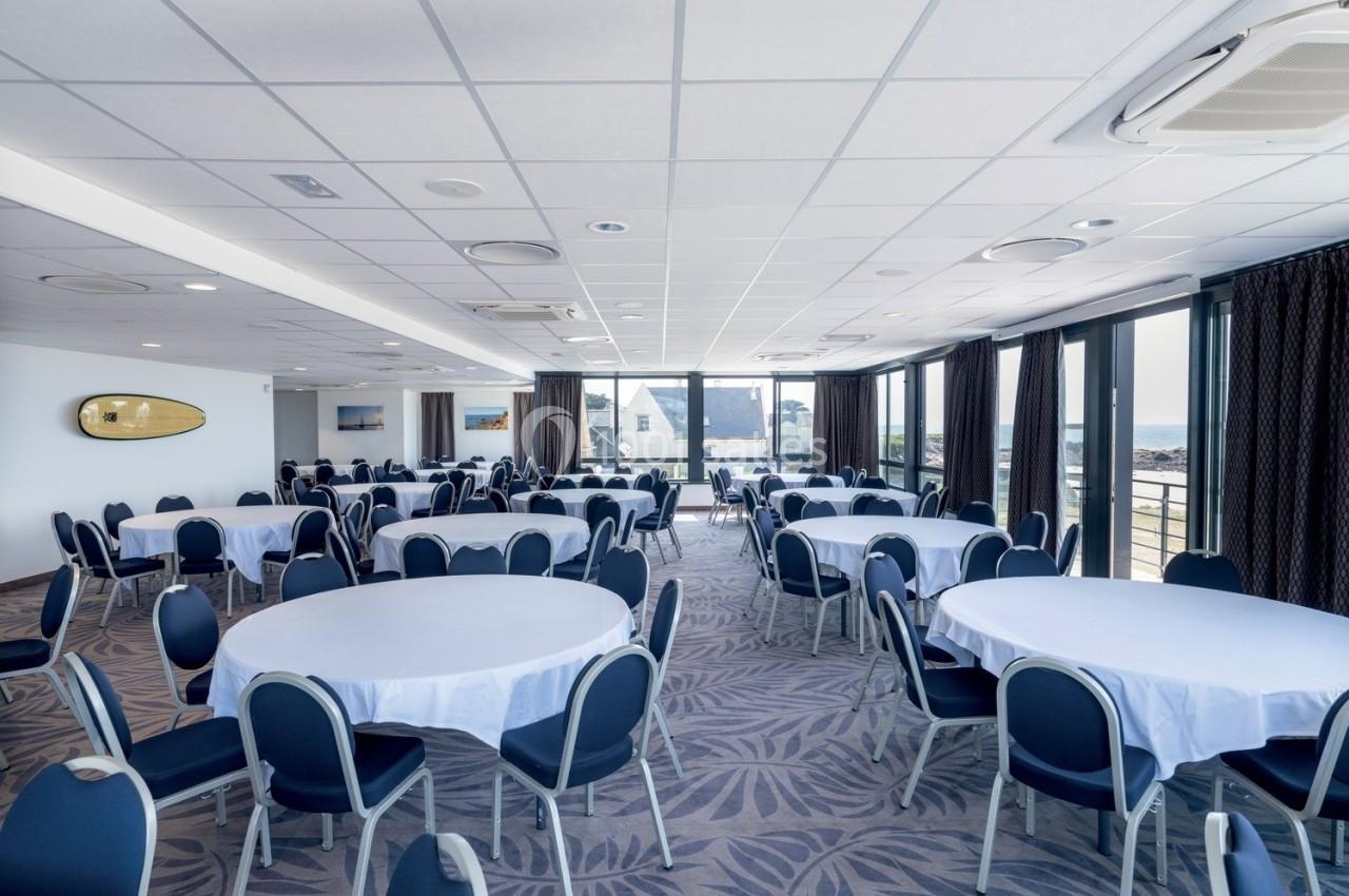Salle lumineuse avec tables rondes dressées de nappes blanches, chaises bleues et grandes baies vitrées donnant sur l…