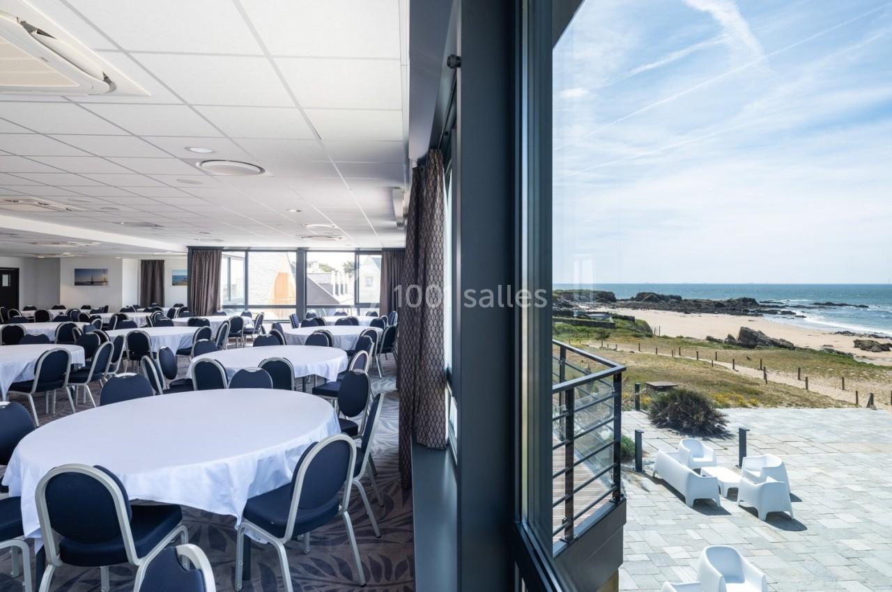 Salle de réception lumineuse avec tables rondes dressées, vue sur une terrasse et la mer à travers de grandes baies vitrées.