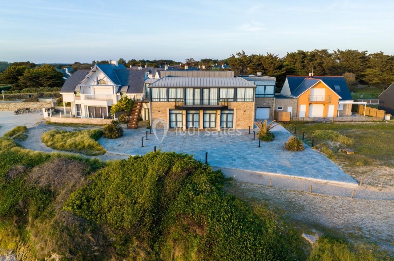 Vue d'une maison moderne en bord de mer entourée de végétation et de chemins sablonneux.
