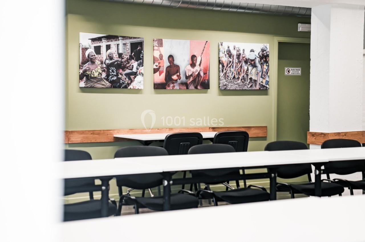 Salle de réunion avec des tables blanches, des chaises noires et des photos accrochées sur un mur vert.