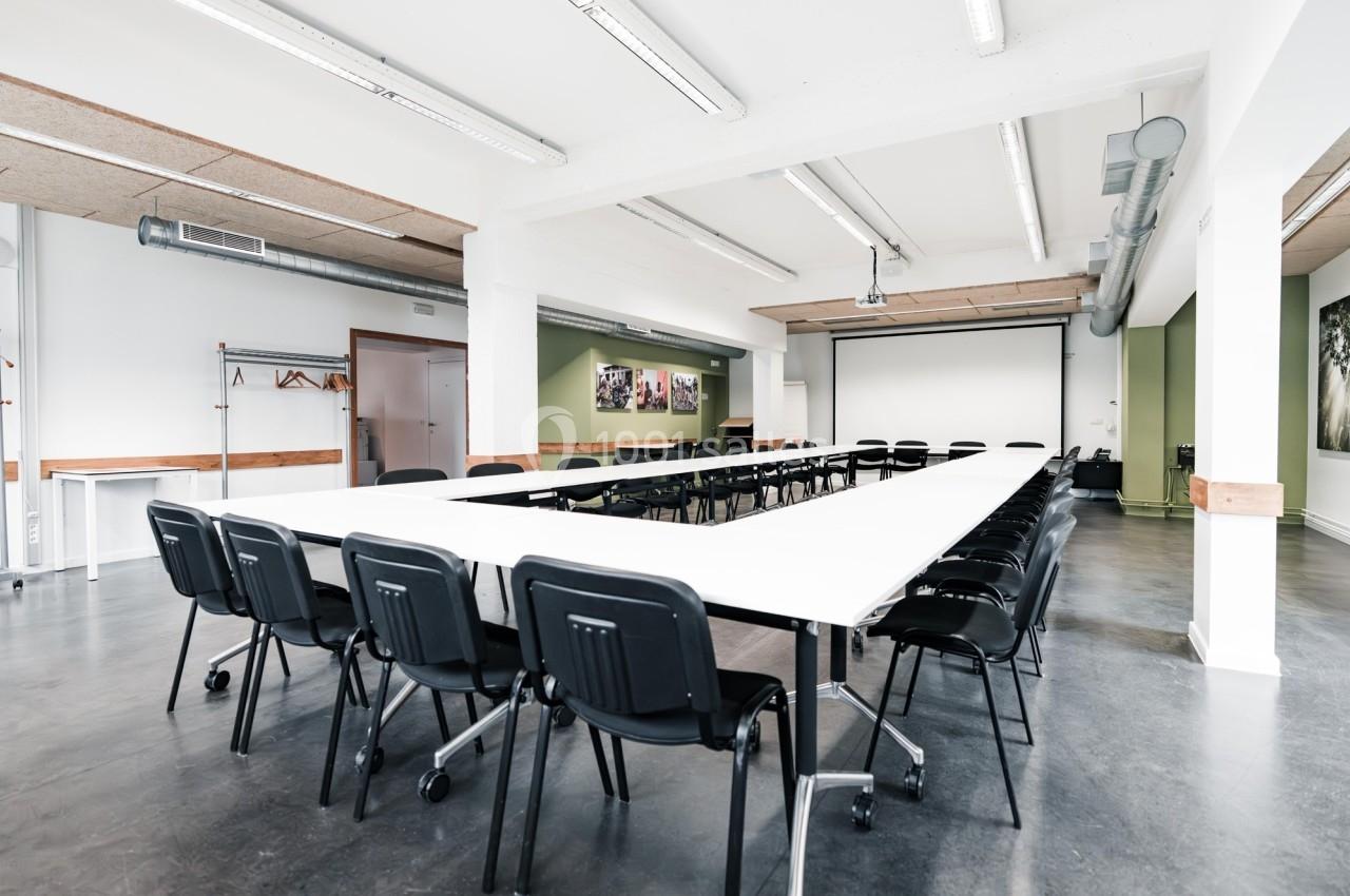 Salle de réunion lumineuse avec tables disposées en U, chaises noires et écran de projection en fond.