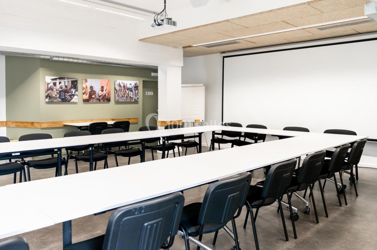 Salle de réunion lumineuse avec tables disposées en U, chaises noires, écran de projection et tableaux au mur.